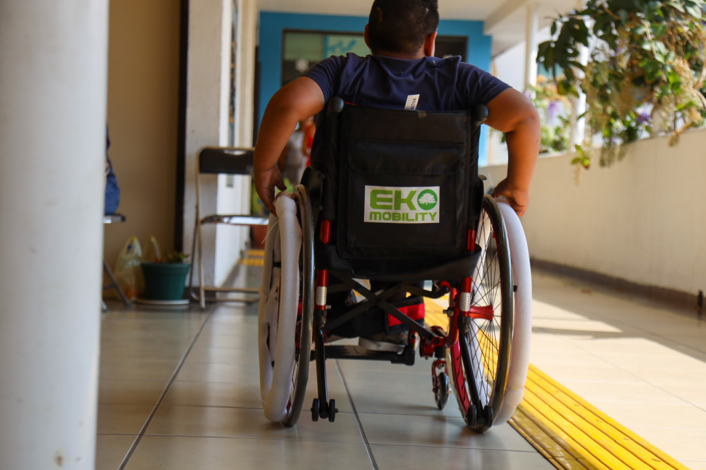 Joven en silla de ruedas activa, de espaldas se impulsa por si solo por un pasillo, al lado de él se enceuntra una guía podotáctil